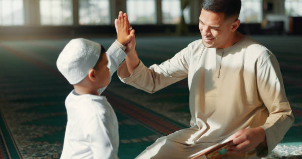 イスラム教徒、モスクでハイタッチをし、父親と息子が信仰、信念、宗教のためにコーランを一緒に勉強します。家族、子供、ラマダン、男の子にイスラムの成功を教える男性 - praying joy indoors lifestyles ストックフォトと画像