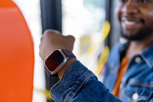 Amidst boarding the bus, a young African American man taps his smartwatch against the fare scanner, modernizing the ticket payment process