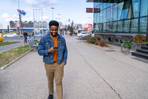 Standing on a street, a young African American man is focused on text messaging, engaging with his phone amid the city's backdrop