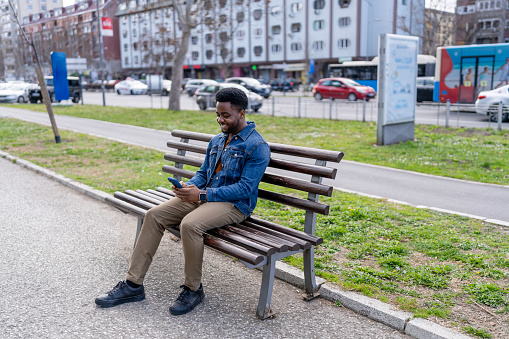 A young African American man finds himself engrossed in his mobile phone while sitting comfortably on a bench, a scene merging public solitude with digital engagement