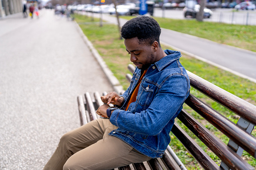 A young African American man looks at his smartwatch while sitting on a bench, checking updates or notifications in a quiet moment outdoors
