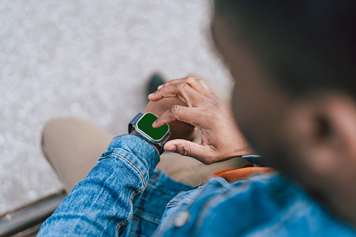 Seated on a bench, a young African American man interacts with his smartwatch, a modern accessory that keeps him connected on the go