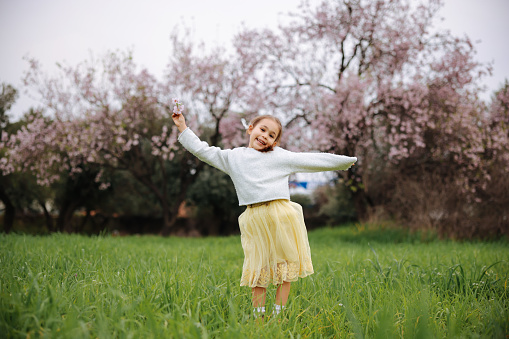 A pretty little girl is picking flowers in a lush meadow at sunset. She's alone in a peaceful natural setting, with a forest in the background. Blooming trees