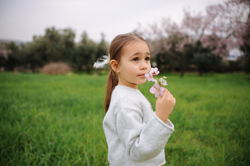 A pretty little girl is picking flowers in a lush meadow at sunset. She's alone in a peaceful natural setting, with a forest in the background. Blooming trees