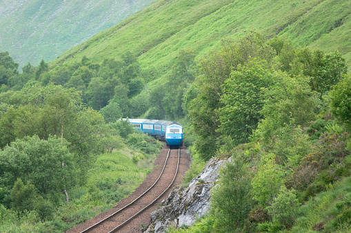 Blue passenger train snakes through the green valleys of the Scottish Highlands on a cloudy day. Comfortable and scenic traveling along the railway tracks that meander through verdant hilly landscape.