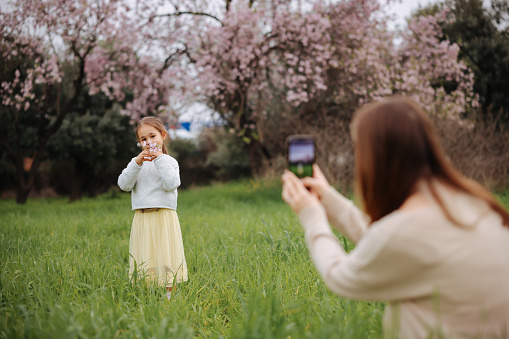 Young mother takes pictures on her daughter’s phone in the flowering garden. Photo shoot for social networks. family walk in the spring garden. Blooming trees