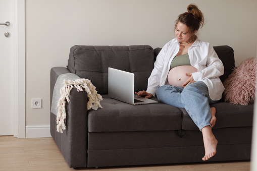 Attractive pregnant young lady working remotely online from home office. Close up woman hands on laptop with big belly advanced pregnancy. selective focus