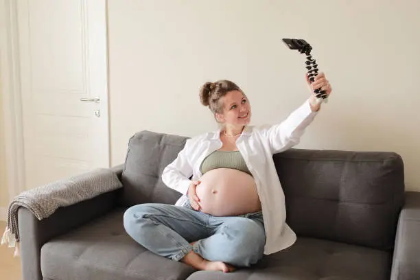 Photo of Happy expecting mother and popular vlogger is recording video for online blog sits on sofa in modern flat and showing her big naked belly looks at smartphone camera. Pregnancy and blogging concept