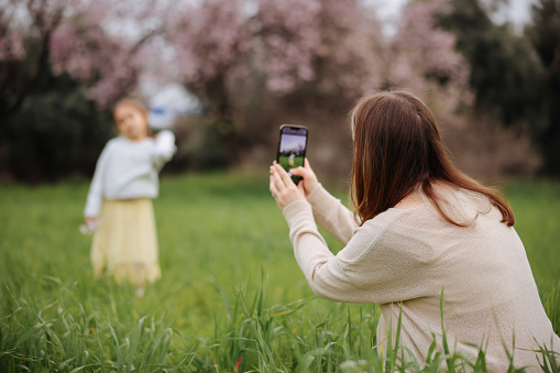 Young mother takes pictures on her daughter’s phone in the flowering garden. Photo shoot for social networks. family walk in the spring garden. Blooming trees