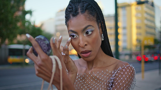 African american model applying makeup at evening street closeup. Afro braids chick woman looking hand mirror correcting her look at sunset avenue. Brown skin lady preparing for date at downtown