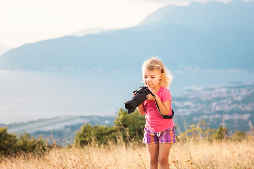 Little kid is playing with camera and taking photo of sea and mountains as master. Blonde girl is traveling photographer.