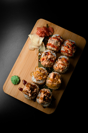 Top view of sushi rolls with sesame, toppings and various sauces lie on a wooden board on a black background isolated