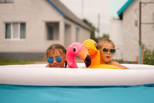 Funny girls in sunglasses swim in a small inflatable swimming pool in the yard next to the house