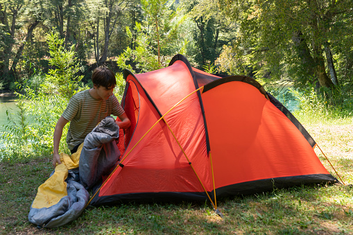 Young man preparing his tent and sleeping bag at a Patagonian campsite surrounded by forests, rivers and mountains during his summer vacation in Argentina.