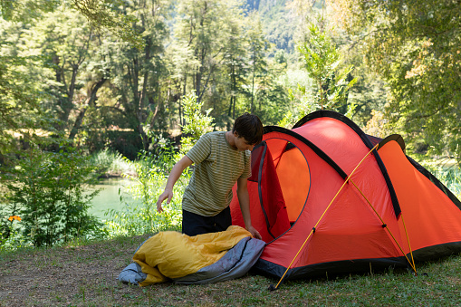 Young man preparing his tent and sleeping bag at a Patagonian campsite surrounded by forests, rivers and mountains during his summer vacation in Argentina.