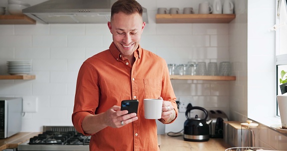 Man, drinking coffee and typing on phone or browsing social media at home, happy and relax on weekend. Male person, communication and mobile app for online conversation, humor and laughing in kitchen