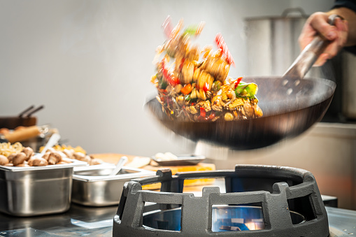 Motion blurred photo of unrecognizable male chef tossing vegetables and meat in the air while stir frying in a wok in a professional kitchen.