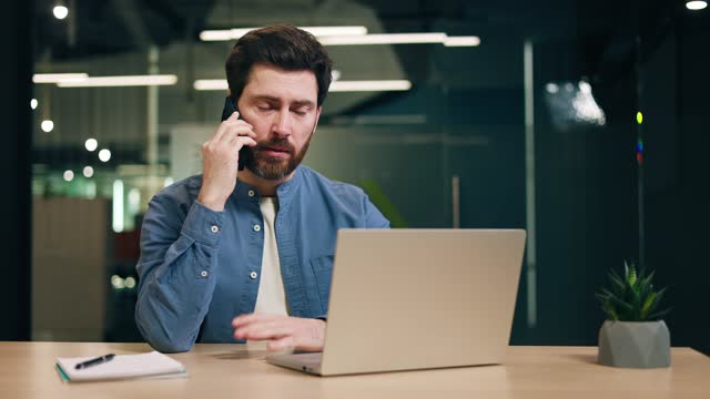Male freelancer speaking on phone while working in comfortable office space. Man wearing informal attire sitting at desktop with laptop and answering questions of employer while gesturing with hand.