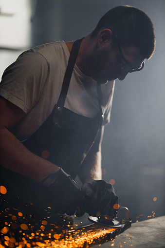 A skilled metalworker in safety glasses and gloves is intensely grinding a piece of metal, with sparks flying around in a dimly lit workshop.