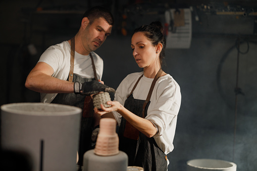 A husband and wife team work collaboratively in their family-owned small workshop, producing beautiful and modern concrete pots for plants, showcasing their passion and craftsmanship.
