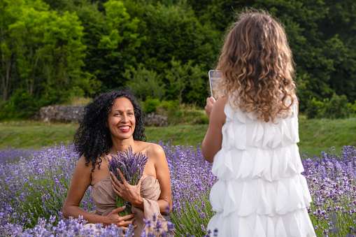 A Latina woman with her daughter holds a bunch of lavender in the middle of a lavender field at sunset. A daughter takes a photo with mobile phone of her mother in a lavender field.