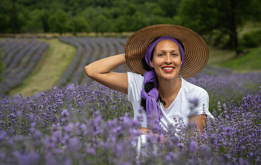 A Latina woman with a purple headscarf and a hat is holding a bunch of lavender in the middle of a lavender field at sunset. She is looking at the camera