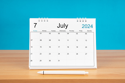 July monthly desk calendar for 2024 year and pen on wooden table with blue color background.