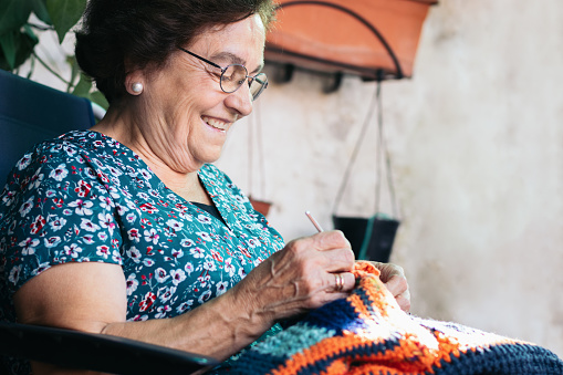 Elderly woman crocheting a blanket. She is sitting in a chair in the patio of her house. She is concentrating on her task.