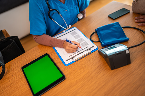 A female healthcare worker in professional attire is indoors, focused on writing patient information on a medical chart. A blood pressure monitor, tablet with green screen, and other medical accessories are visible on the desk.