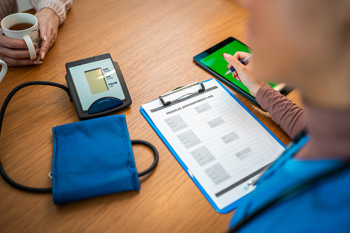 Mixed race nurse checking patients blood pressure with a digital sphygmomanometer. She is seated indoors, casually dressed, with health monitoring equipment and medical paperwork on the table.