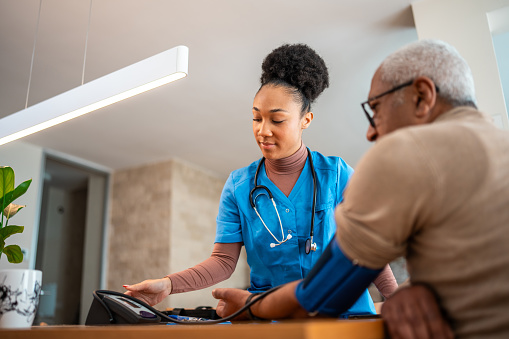 Black female health professional in blue scrubs assists an elderly black male patient, offering care and guidance. They are indoors, interacting over a blood pressure monitor, depicting a clinical or medical consultation scene.