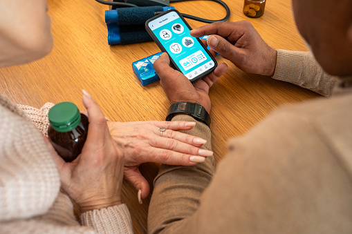 Close-up view of elderly Caucasian woman's hands guiding a senior Black man through a healthcare application on a smartphone, indoors, with medication and blood pressure monitor nearby. Depicts technology usage for health management at home.