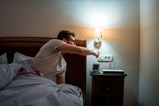 A young Caucasian male in casual nightwear prepares for sleep by switching off the bedside lamp. The image is indoors, highlighting a nighttime routine.
