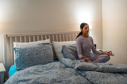 An Indian female in serene meditation on her bed, practicing a bedtime routine in a peaceful bedroom setting. She wears comfortable sleepwear and performs a seated yoga pose to promote relaxation.