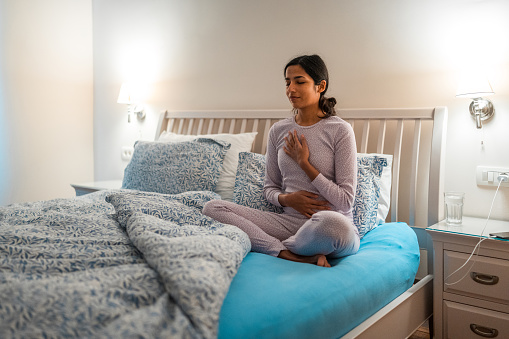 An Indian female in comfortable sleepwear sitting on a bed practicing yoga, with a serene expression indoors. Bedroom setting demonstrates a calming nightly routine.