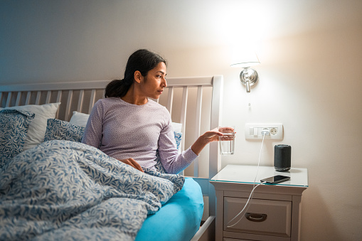 A serene Indian female in casual nightwear sitting on her bed, holding a glass of water, with a lit bedside lamp in a home interior at night, encapsulating a nightly hydration routine.