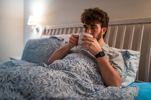 Caucasian male in casual nightwear, holding a cup, lying in bed unable to sleep, depicting insomnia indoors at night with a concerned expression.