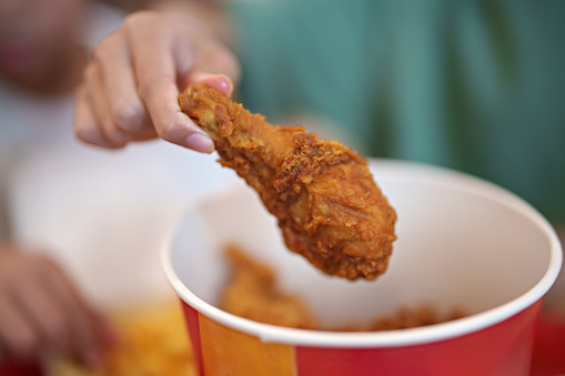 In the cropped image, a little boy's hand is seen taking a fried chicken drumstick from a bucket.