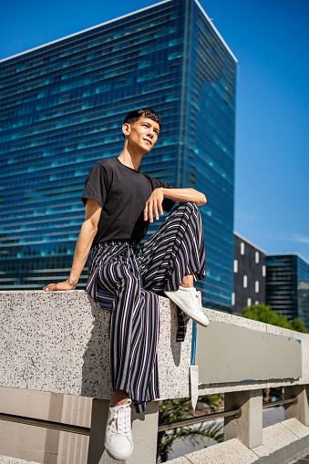 Young model wearing stylish pants sitting on a cement railing outside in the city in summer