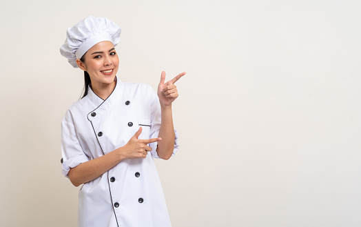 Smiling Young beautiful asian woman chef in uniform pointing finger to copy space isolated background. Cooking woman Occupation chef or baker People in kitchen restaurant and hotel.