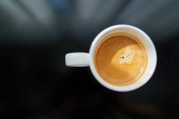 From a directly above view, a hot espresso with thick crema is presented in a white coffee cup, placed on a black glass table at a cafe.