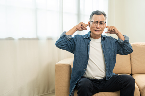 Handsome asian mature old man stressed and frustrated covering his ears with his hands due to loud noise Neighbors disturb around the home. Annoyed noise sound pollution. Asian old man blocking ears
