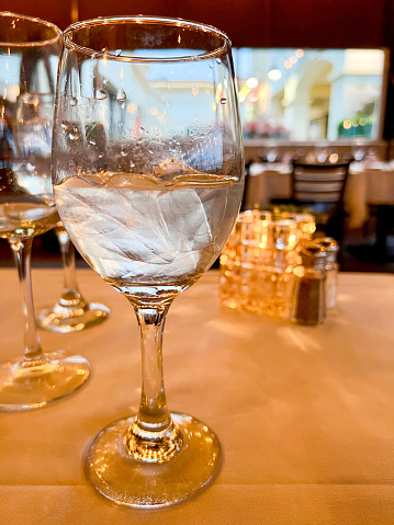 Close up of a wine glass with ice water along with two other wine glasses, table candle, salt and pepper behind it at a restaurant.