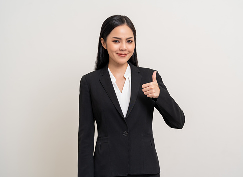 Attractive Young asian business woman thumbs up looking to camera standing pose on isolated white background. Latin Female around 25 in black suit portrait shot in studio.