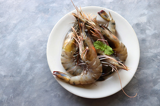 Fresh shrimp or black tiger prawns. Uncooked on a white ceramic plate with coriander and mint leaves. on a moody background with copy space. Kolambi in Marathi.