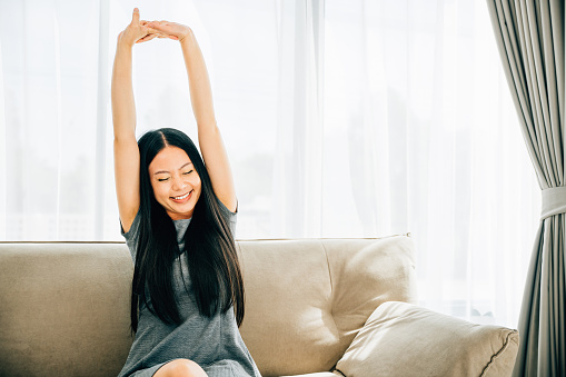 Happy woman on sofa raises arms stretches after sitting for long time. Lifestyle of a cheerful businesswoman in a luxury living room. Wellbeing joy and carefree relaxation.