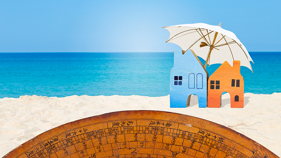 Miniature house with beach umbrella and old wooden Chinese fengshui compass on the beach, summer outdoor day light, property industry, real estate business