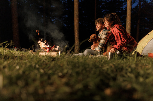 Young happy couple and their dog relaxing by the fire during camping night in nature. Copy space.