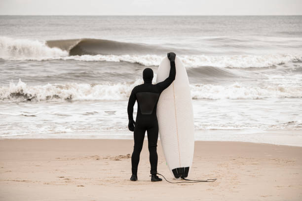 Sender male surfer stands on the seashore and holds his surfboard against the background of the choppy sea