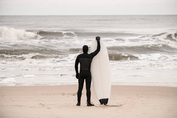 Slim male surfer in black wetsuit standing on the seashore and holding his surfboard admiring the sea with waves
